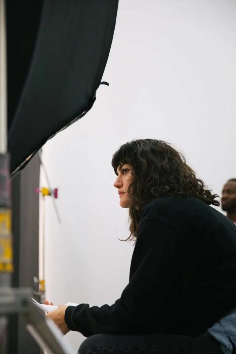 A woman with curly dark hair sitting and looking forward in a studio or classroom, with a white wall in the background and another person slightly visible behind her.