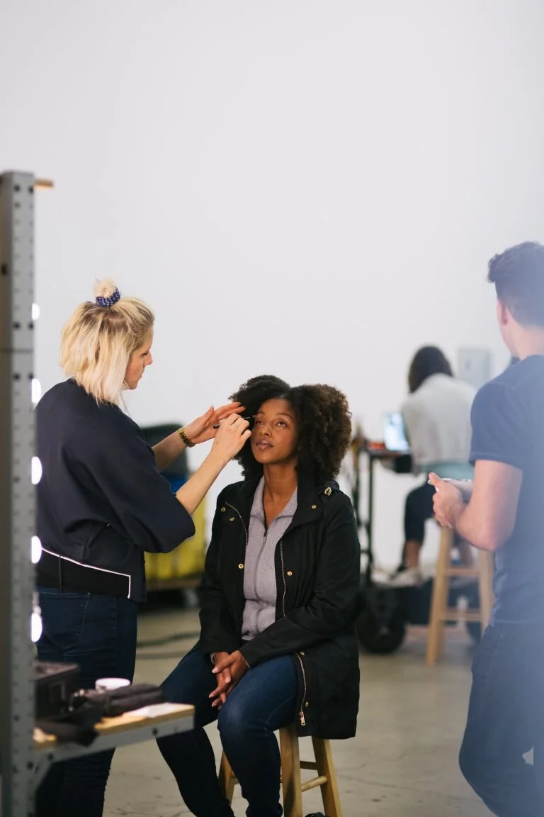 Makeup artist applying makeup to a seated model in a studio setting, with other people working in the background.