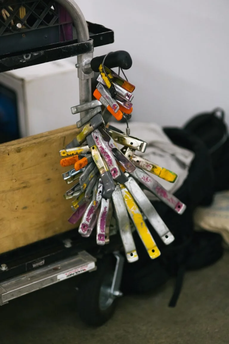 A cluster of paint scrapers hanging from a hook on a tool cart, with a black backpack and other objects in the background.