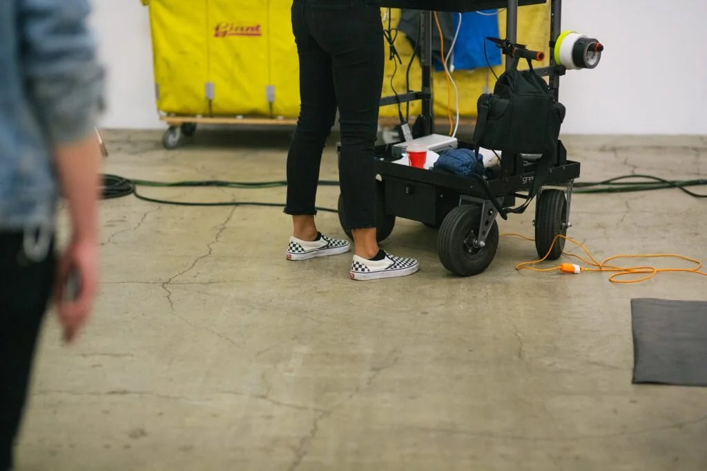 Person wearing checkered slip-on shoes standing next to a black rolling cart with equipment, in an industrial-style space.