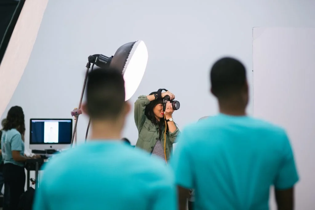 Photographer taking pictures of two men in a studio setting with photography equipment and a computer in the background.