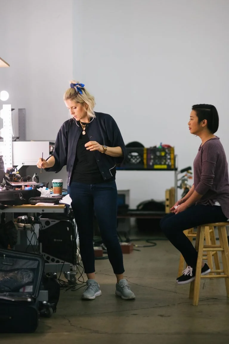 Two women in a photography studio. One standing, working on makeup or a hair touch-up. The other sitting on a wooden stool, waiting. Makeup and photography equipment are visible.