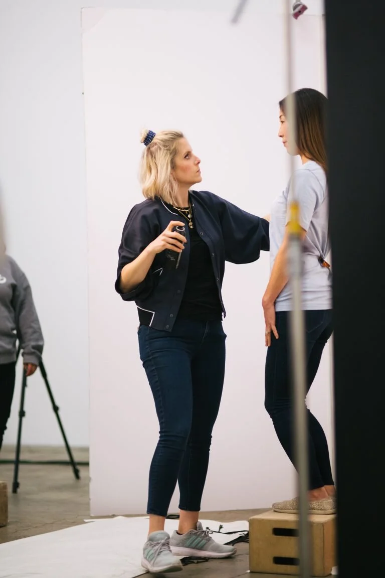 A photographer directs a photoshoot in a studio, with a woman standing on a wooden box posing for the camera, while another woman assists with styling or makeup.