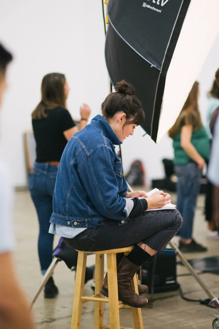 A young woman with dark hair in a bun, wearing a denim jacket, black pants, and brown boots, sitting on a wooden stool, writing or drawing on a notepad in a crowded indoor setting with large lighting equipment overhead and other people standing around.