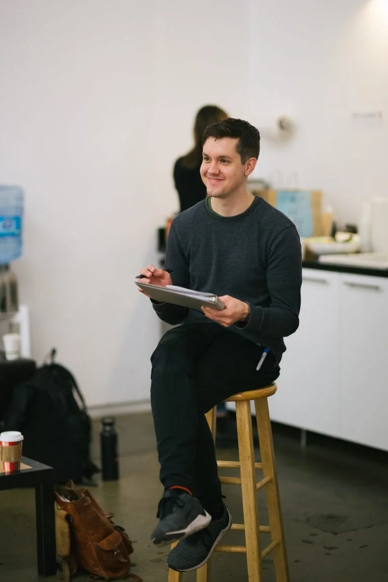 A young man sitting on a wooden stool in an office or break room, smiling while holding a clipboard. In the background, a woman is standing near a counter with various items, and a water dispenser is visible on the left side of the image.