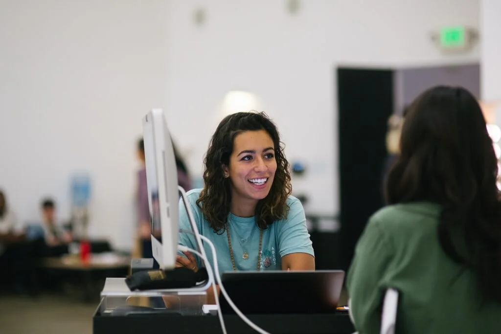 A woman with curly brown hair smiling and talking to another woman with long dark hair at a desk in an office setting.
