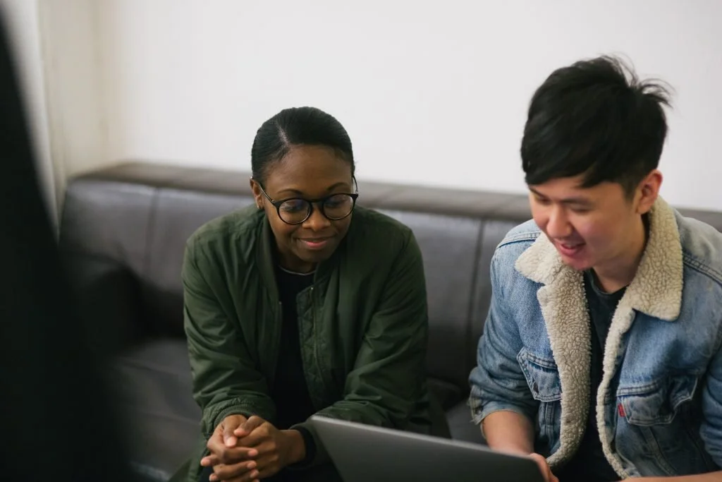 Two young people sitting on a couch looking at a laptop, smiling, indoors.