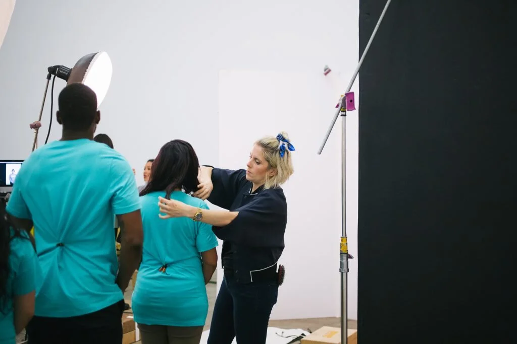 A woman in black scrubs adjusting a girl's hair during a photo shoot with studio lighting and a black backdrop.