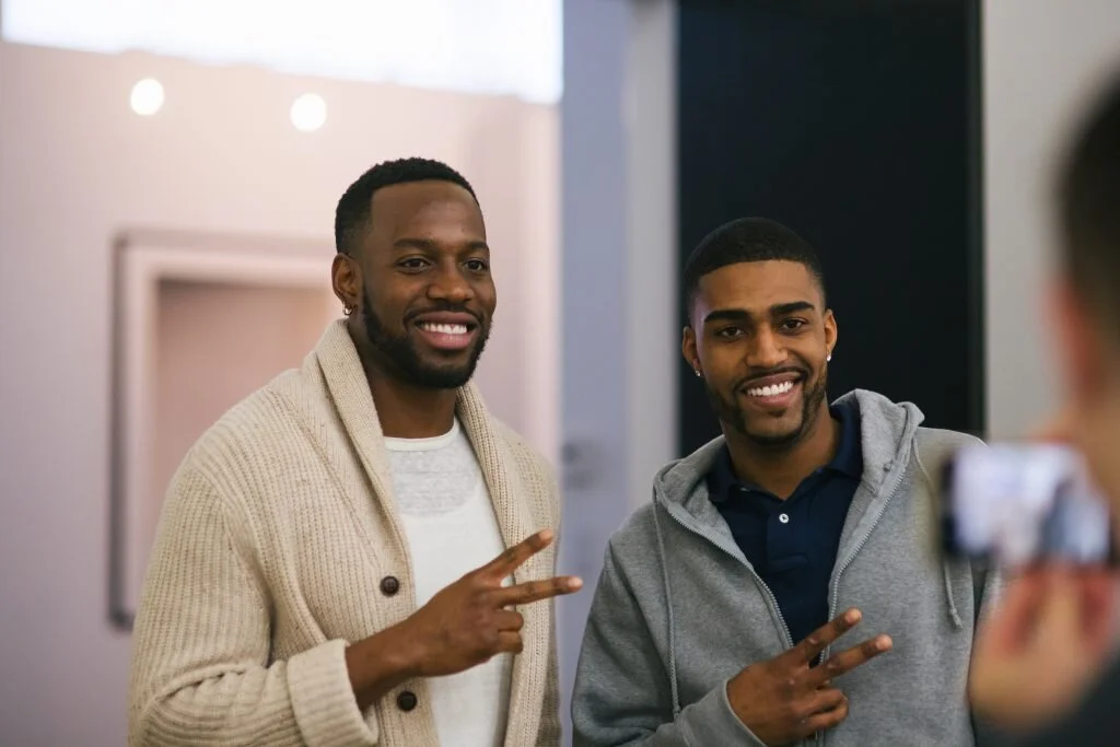 Two young men posing for a photo, smiling, making peace signs with their fingers in an indoor setting.