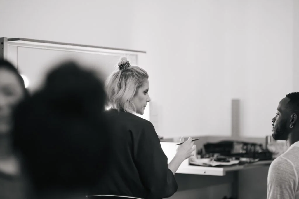 A woman with blonde hair tied up with a scrunchie sitting at a desk, looking at a man with short hair, who is seated facing her in a classroom or office setting.