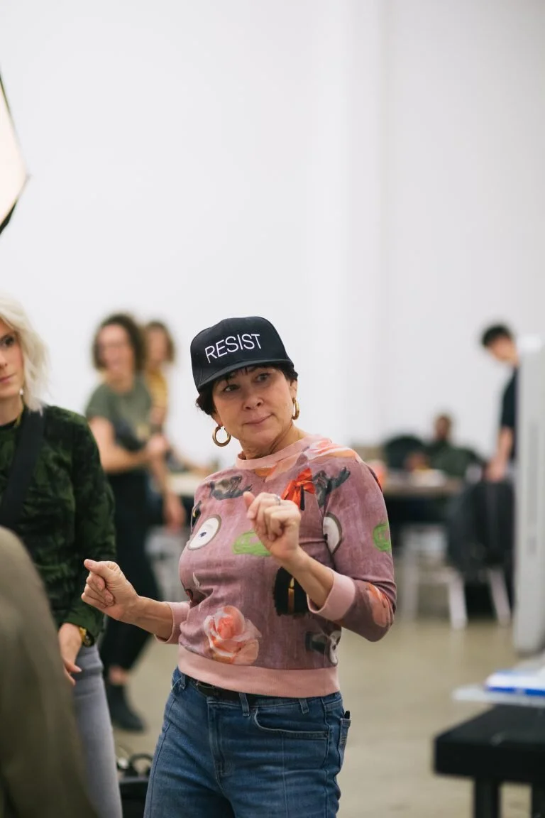 A woman wearing a black cap with the word 'RESIST' in white letters, a colorful patterned shirt, and blue jeans, dancing or gesturing in a room with other people in the background.