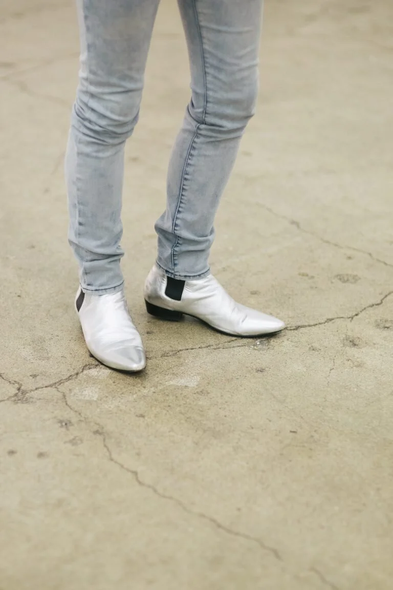 Person wearing light gray jeans and shiny silver pointed-toe cowboy boots standing on a cracked concrete floor.