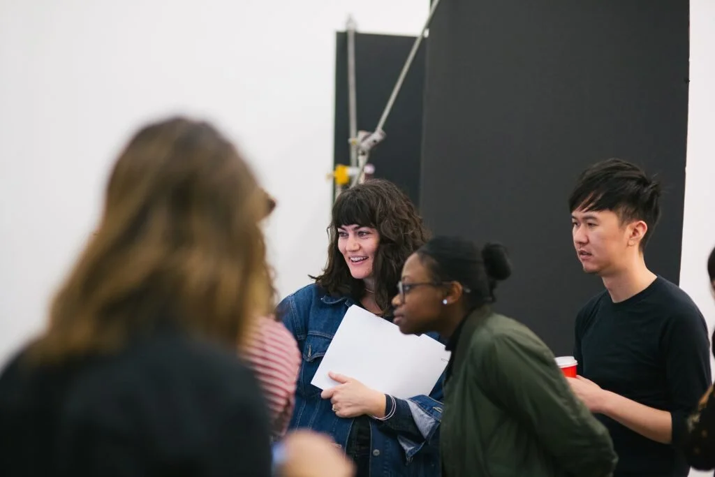 Group of young people socializing indoors, smiling and holding drinks, with a dark wall in the background.