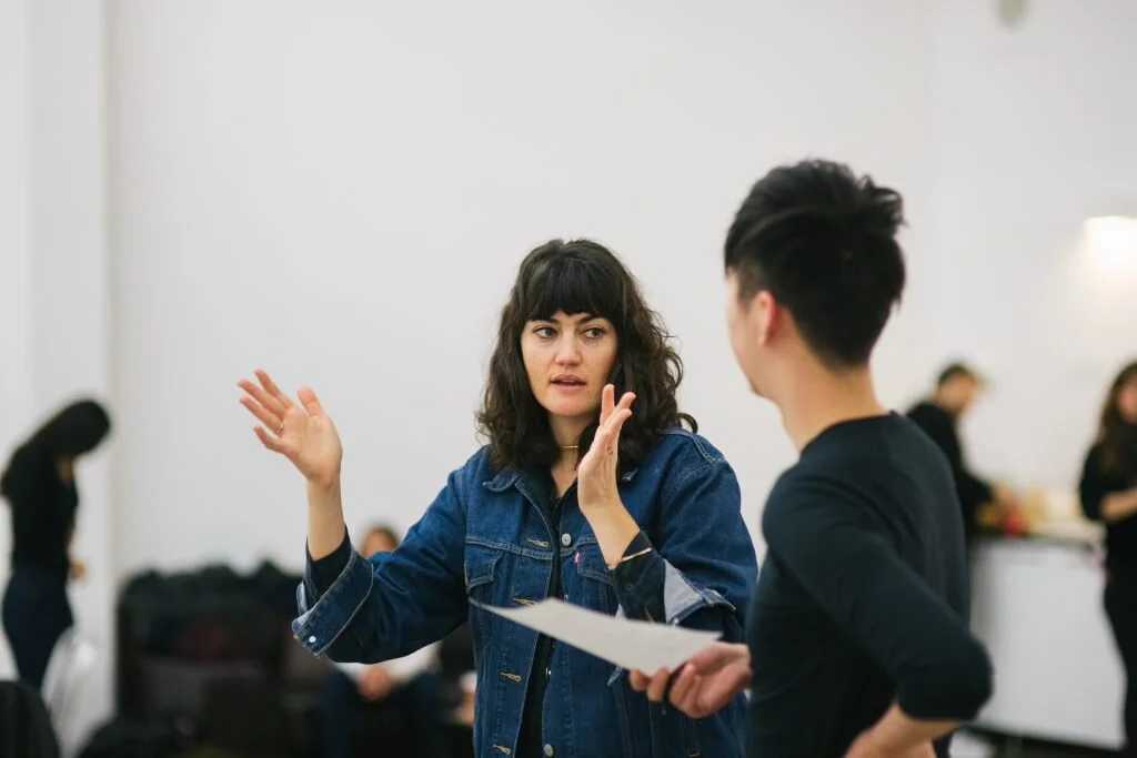 Two women engaging in conversation in an indoor setting with other people in the background