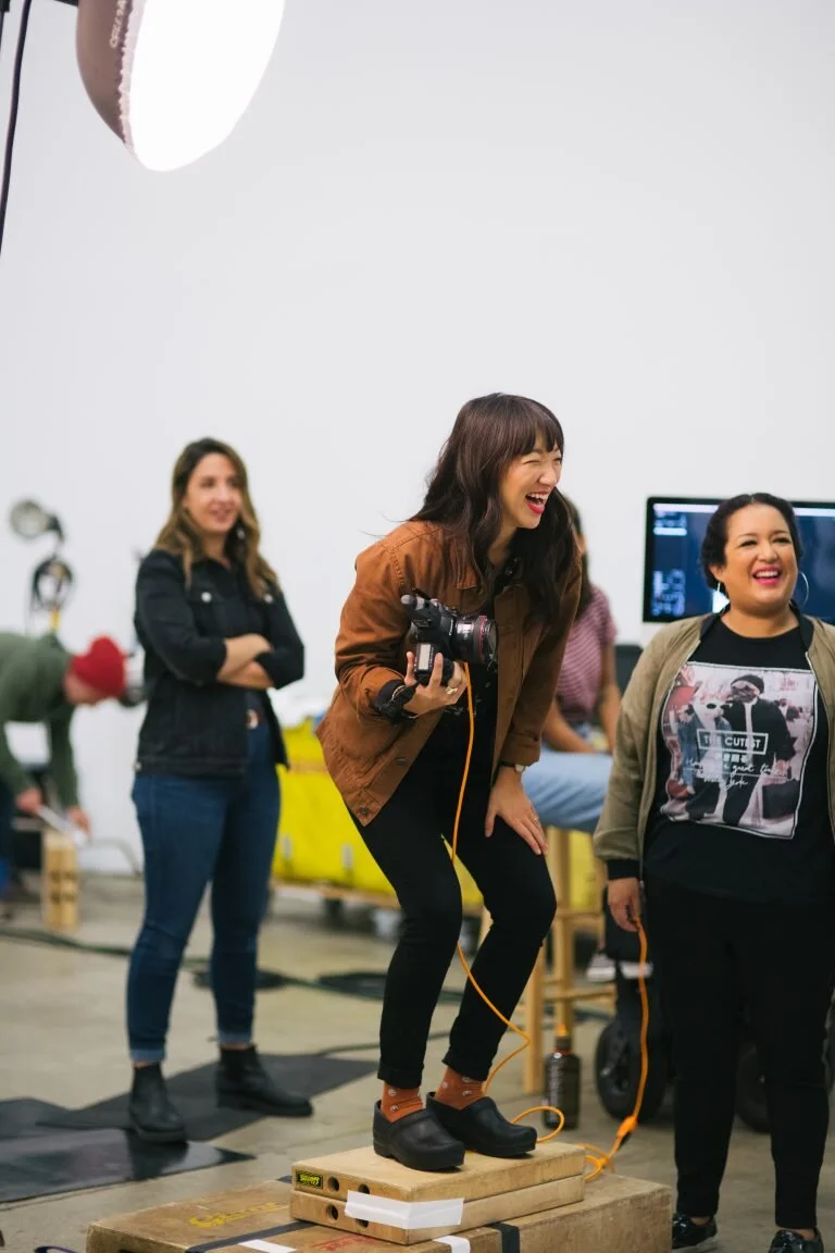 Group of women in a studio environment, with one woman laughing and standing on a wooden plank, holding a camera, while others watch and smile.