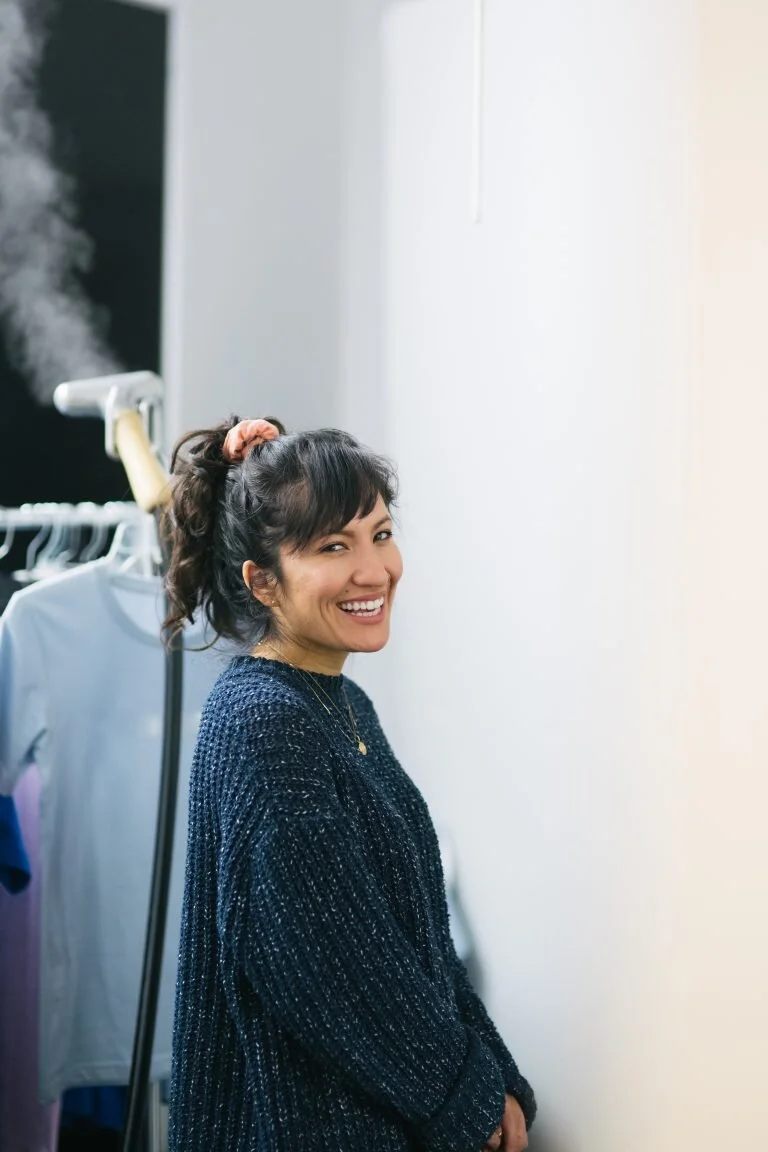 A woman smiling and standing next to a clothing rack in a bright, indoor setting.