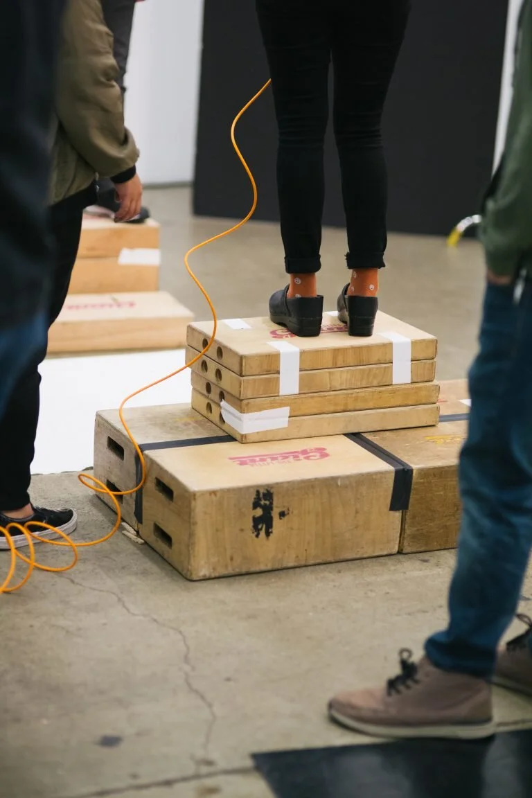 Person standing on a stack of wooden boxes during a fitness or balance test, surrounded by others in an indoor setting.