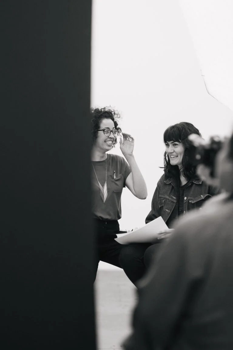 Two women laughing and talking during a photo shoot in a studio, with one holding papers, seen through a partial view of a person with a camera in the foreground.