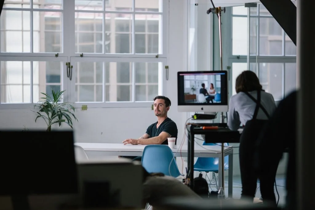 A man sitting in a brightly lit office space with large windows, facing the camera, while a woman in professional attire sits at a desk with a computer displaying the scene.