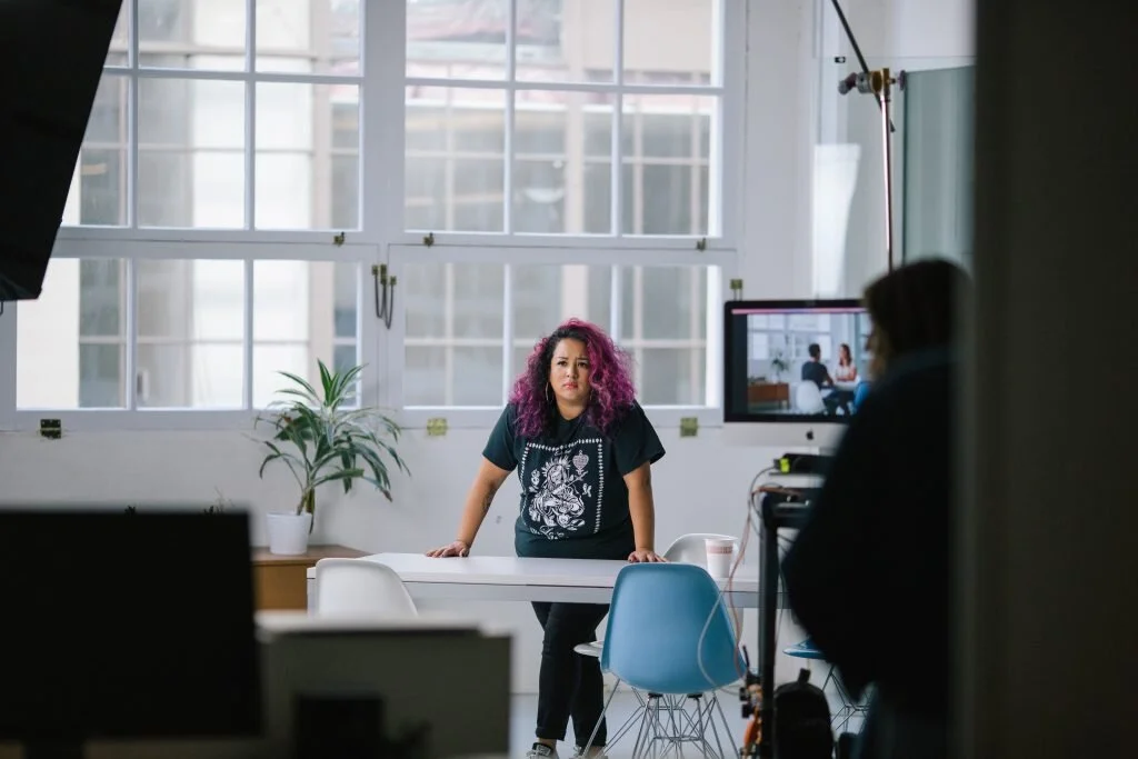 Woman with curly pink hair standing behind a table in a well-lit room, being filmed for a video or interview, with a camera operator and studio equipment visible.
