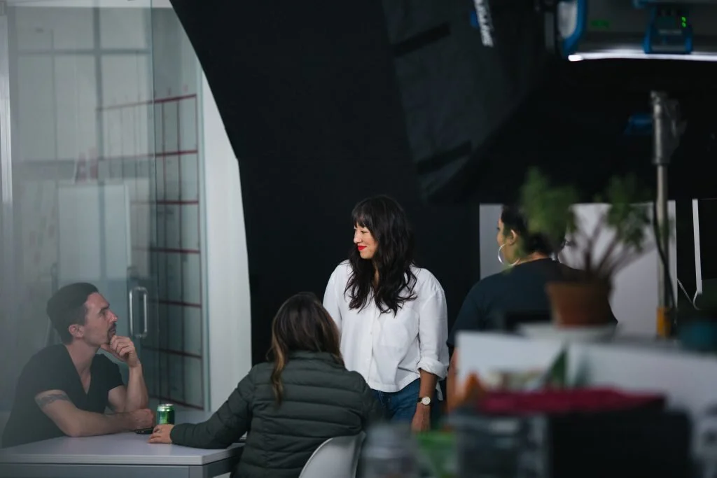 People conversing in a modern indoor space, with a woman standing and three others seated around a table, one holding a beverage.