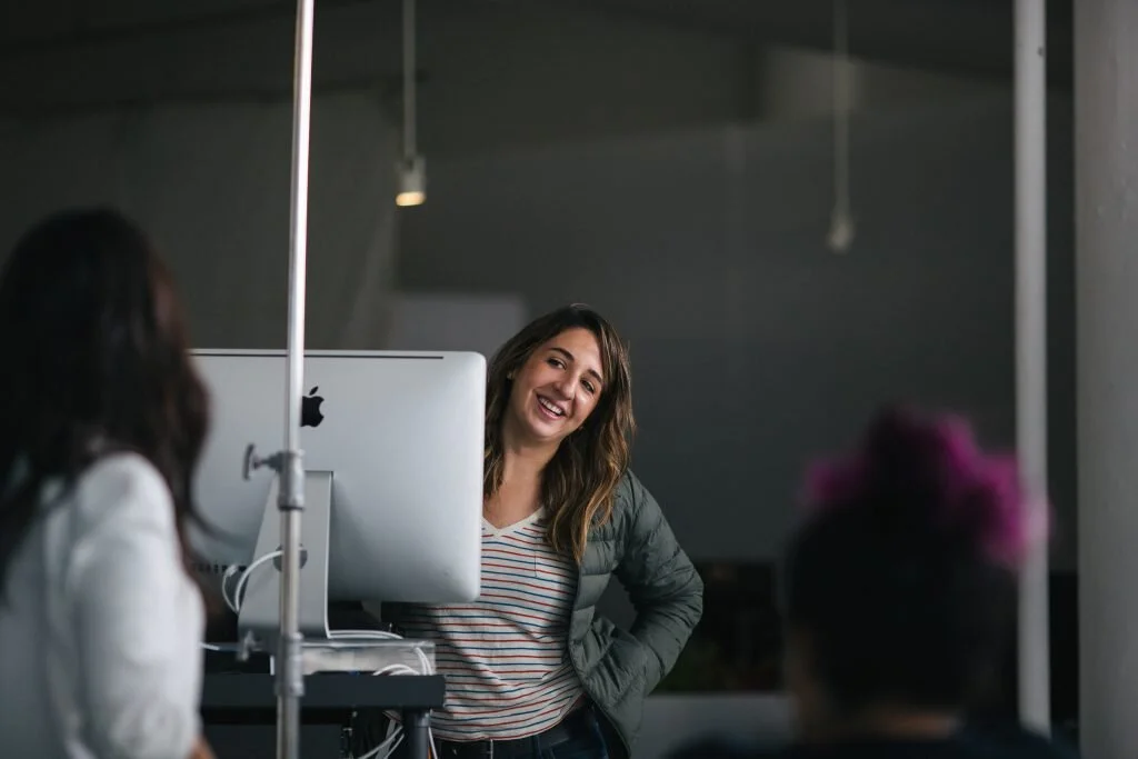 Woman smiling and talking to two others in a room with a computer.