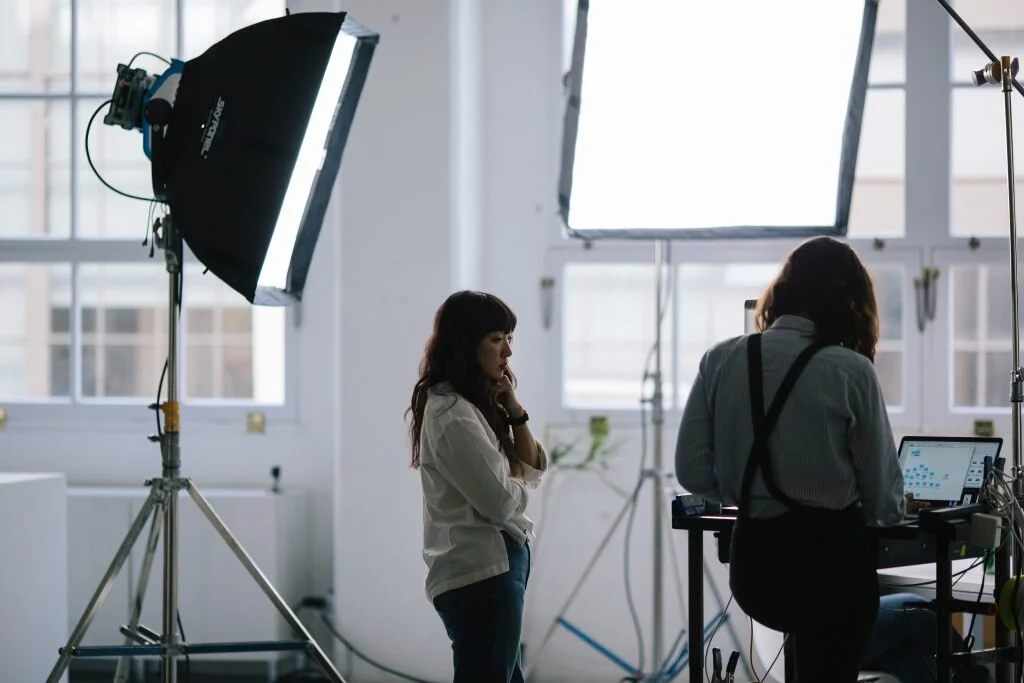 A woman in a white shirt with dark hair looking thoughtful in a photography studio with professional lighting equipment and a person working on a computer.
