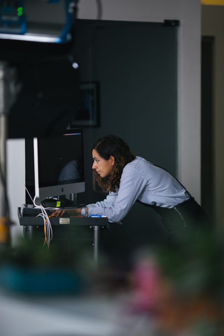 A woman with dark hair leaning over a desk, working on a computer in an office environment.