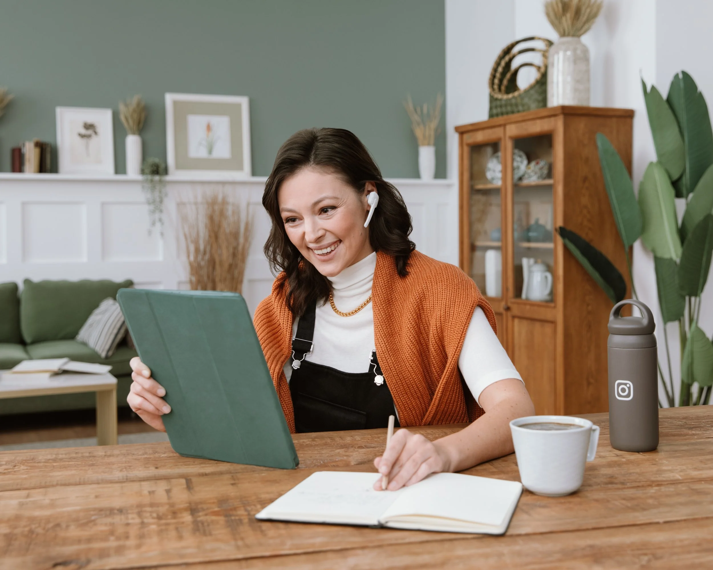 A woman with dark hair wearing an orange sweater and a headset, sitting at a wooden table with an open notebook, a cup of coffee, and a water bottle. She is smiling and looking at a green tablet.