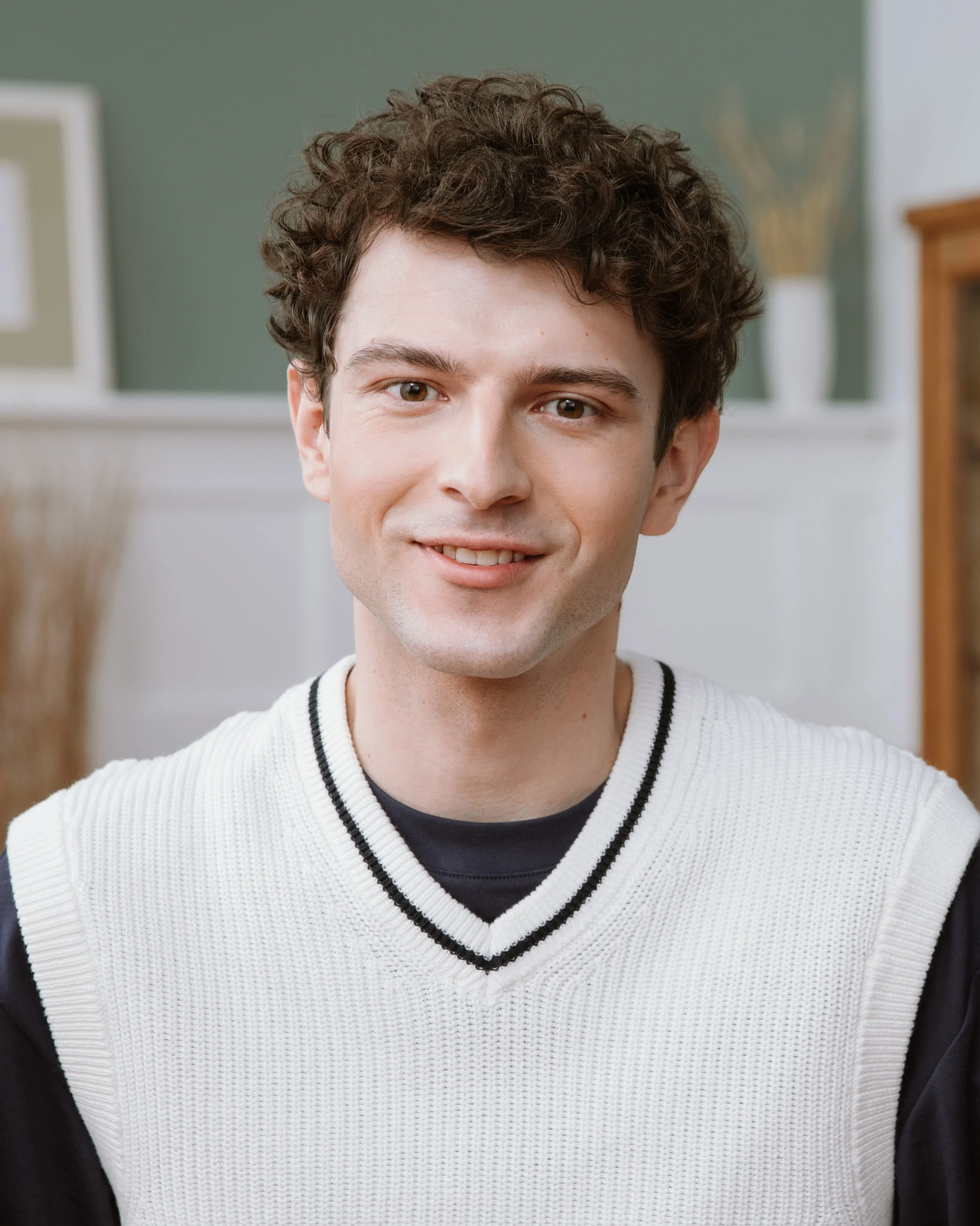 Close-up of a smiling young man with curly brown hair, wearing a white sweater with black trim, indoors.