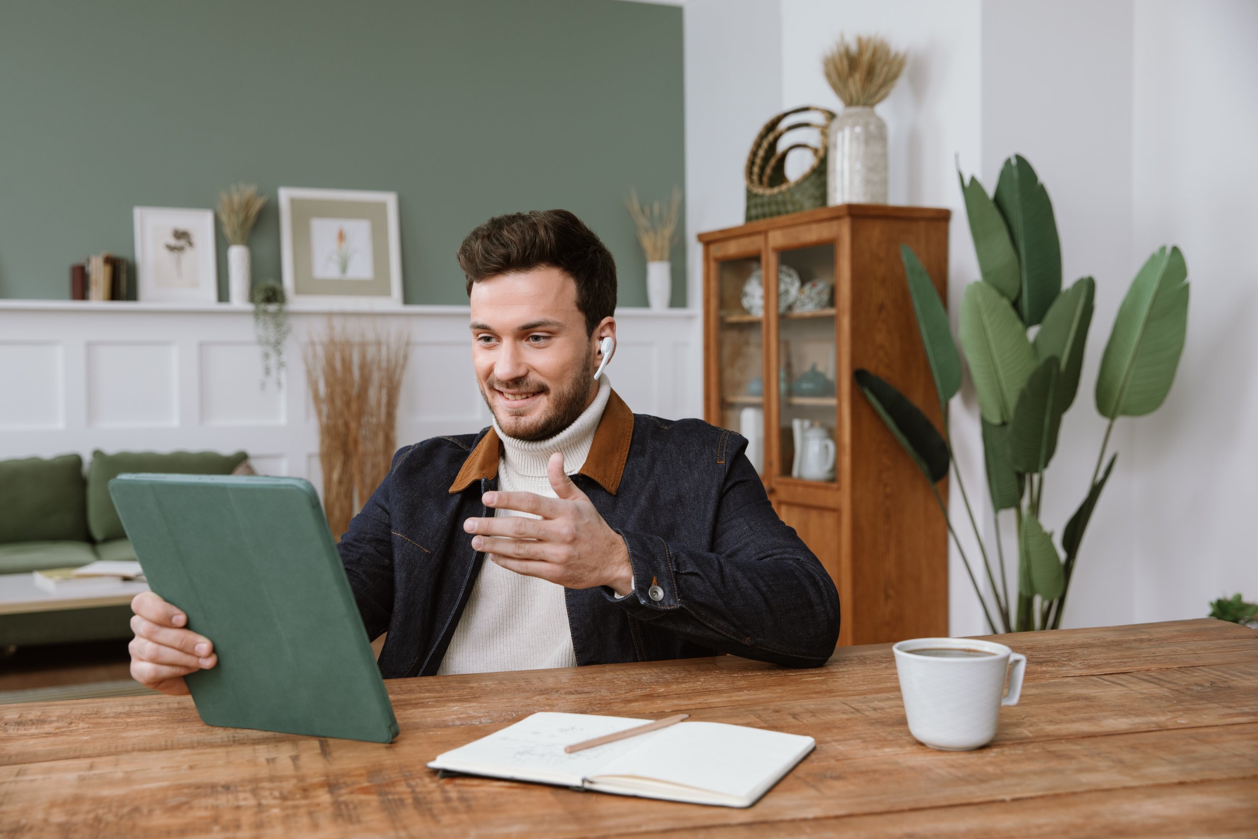 A man with short dark hair and a beard, smiling and wearing a beige turtleneck and a dark jacket, sitting at a wooden table with a green laptop, notepad with a pencil, and a cup of coffee, in a cozy living room with green and white walls, decorative 