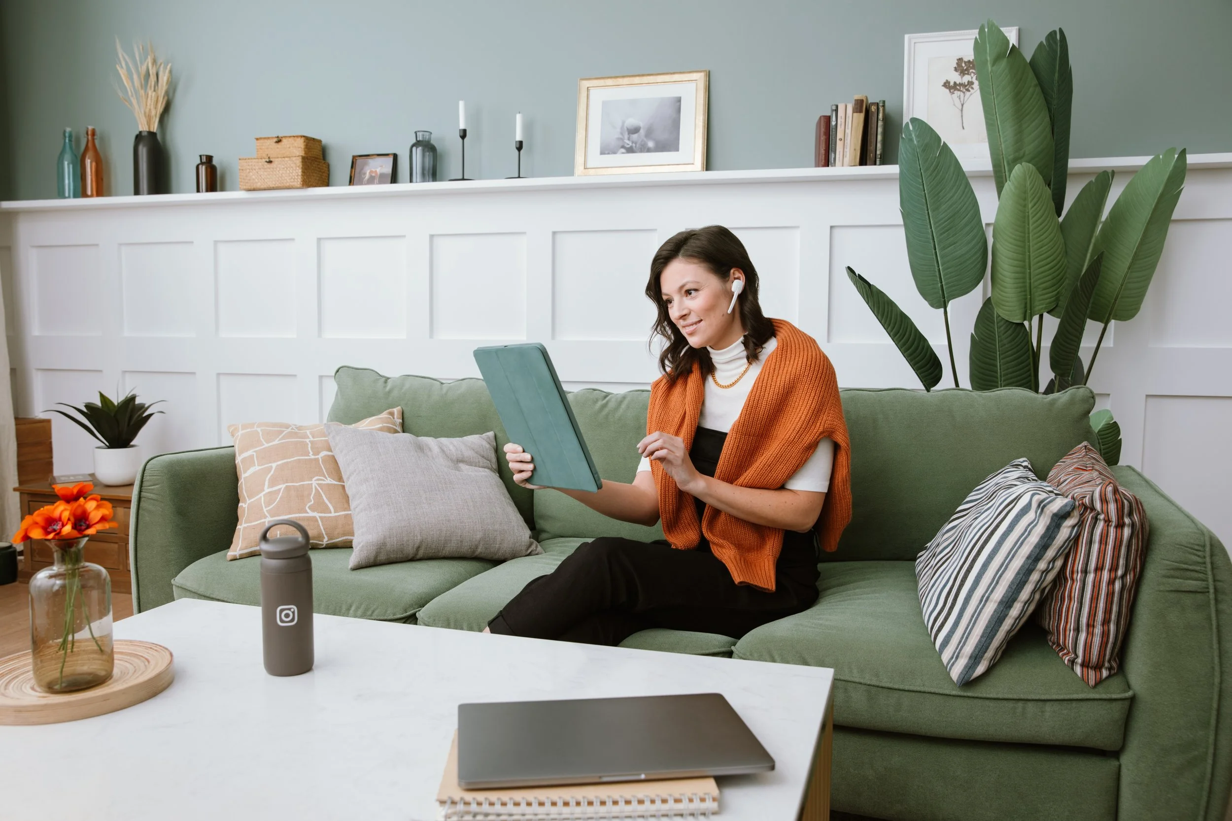 Woman sitting on a green sofa, wearing a white turtleneck and orange vest, with a wireless headset, looking at a tablet. The living room has a white wall with framed pictures, a large plant, and decorative items on the shelf. A white coffee table in 