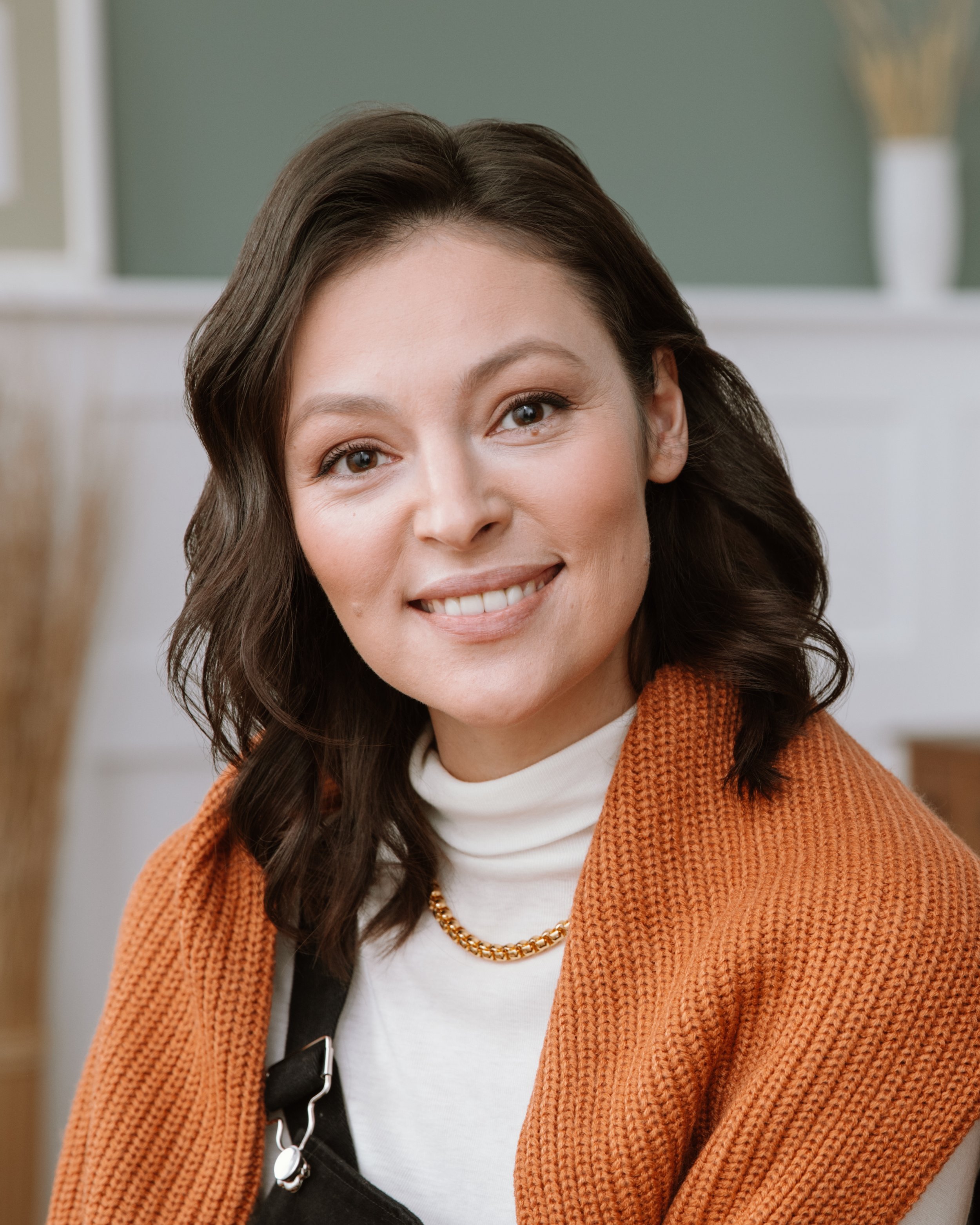 Close-up of a smiling woman with brown hair, wearing a white turtleneck, an orange knitted cardigan, a gold chain necklace, and a black strap bag, in a well-lit indoor setting.