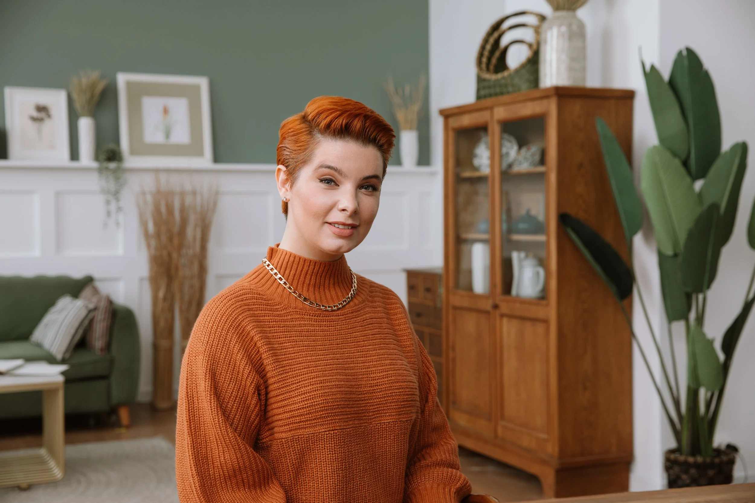 A woman with short red hair wearing an orange sweater and a silver chain necklace, smiling in a cozy living room with green and white walls, decorated with framed pictures, plants, and wooden furniture.