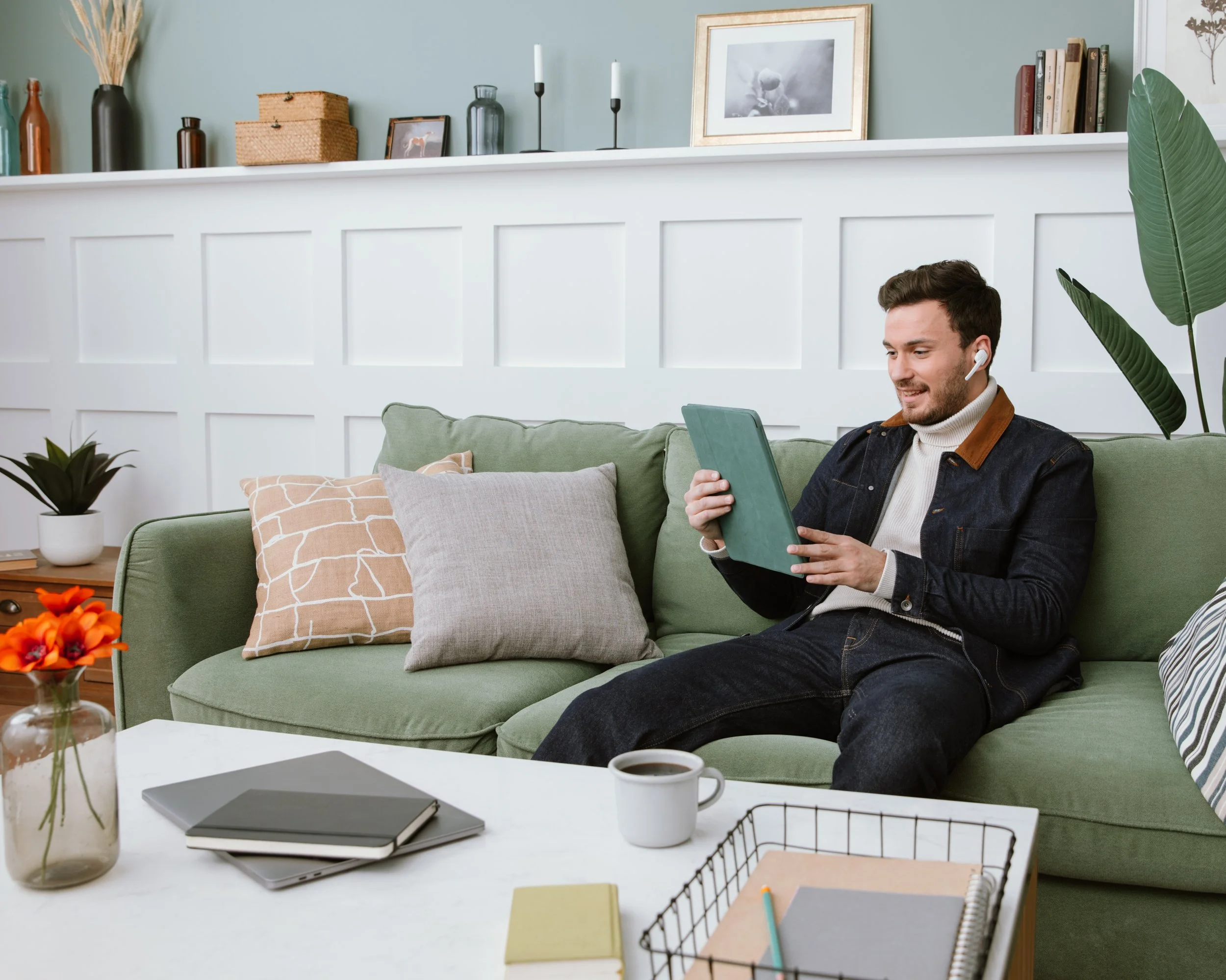 A man with short brown hair wearing a white turtleneck and denim jacket sitting on a green sofa, using a tablet, with a work desk nearby that has a coffee mug, notebook, laptop, and colorful sticky notes, in a brightly lit living room with decorative