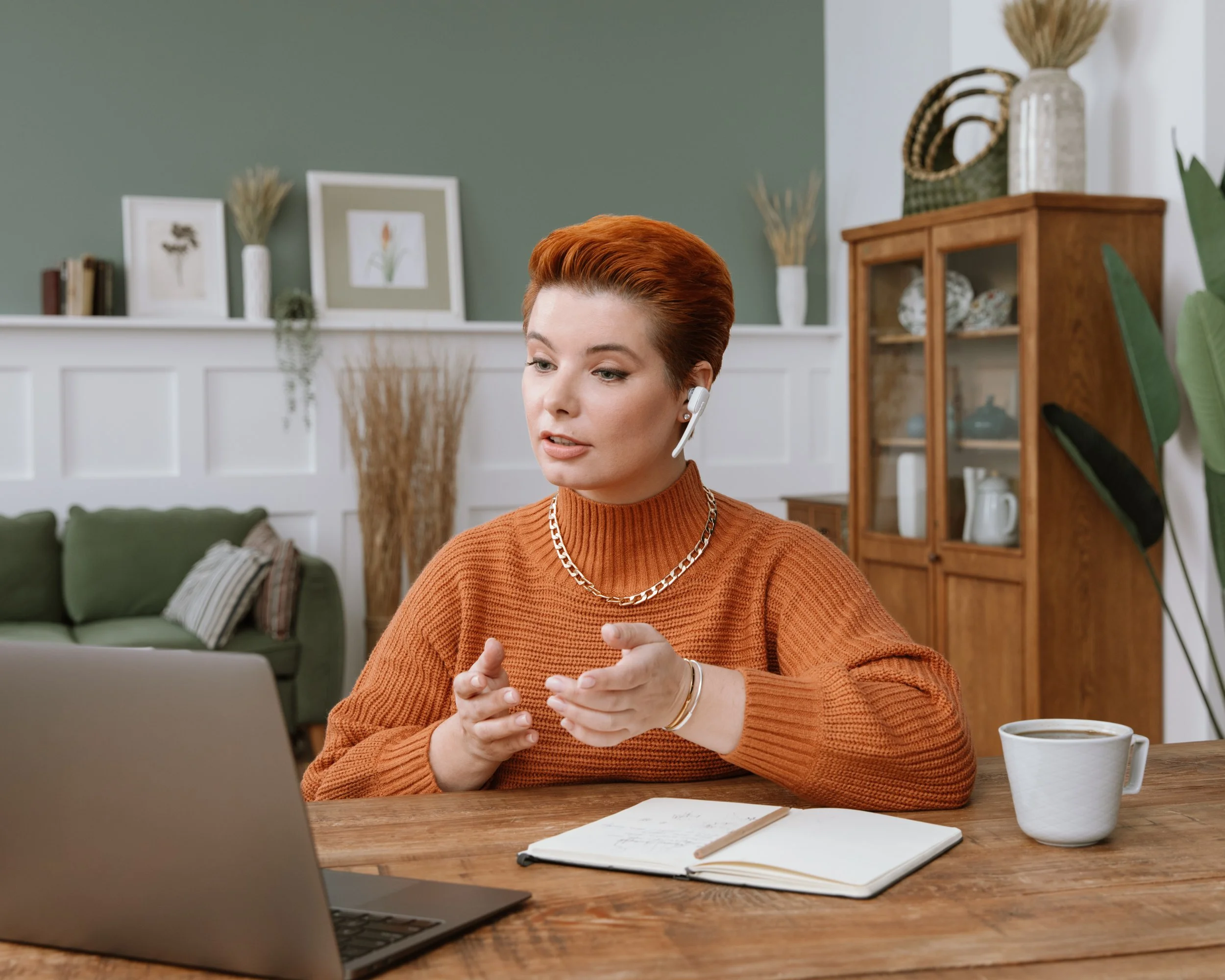 A woman with short red hair and a tan sweater sitting at a wooden table, talking on a headset, with a notebook and pen in front of her, and a white mug to her right, in a room with green and white walls, decorative plants, framed pictures, and a wood