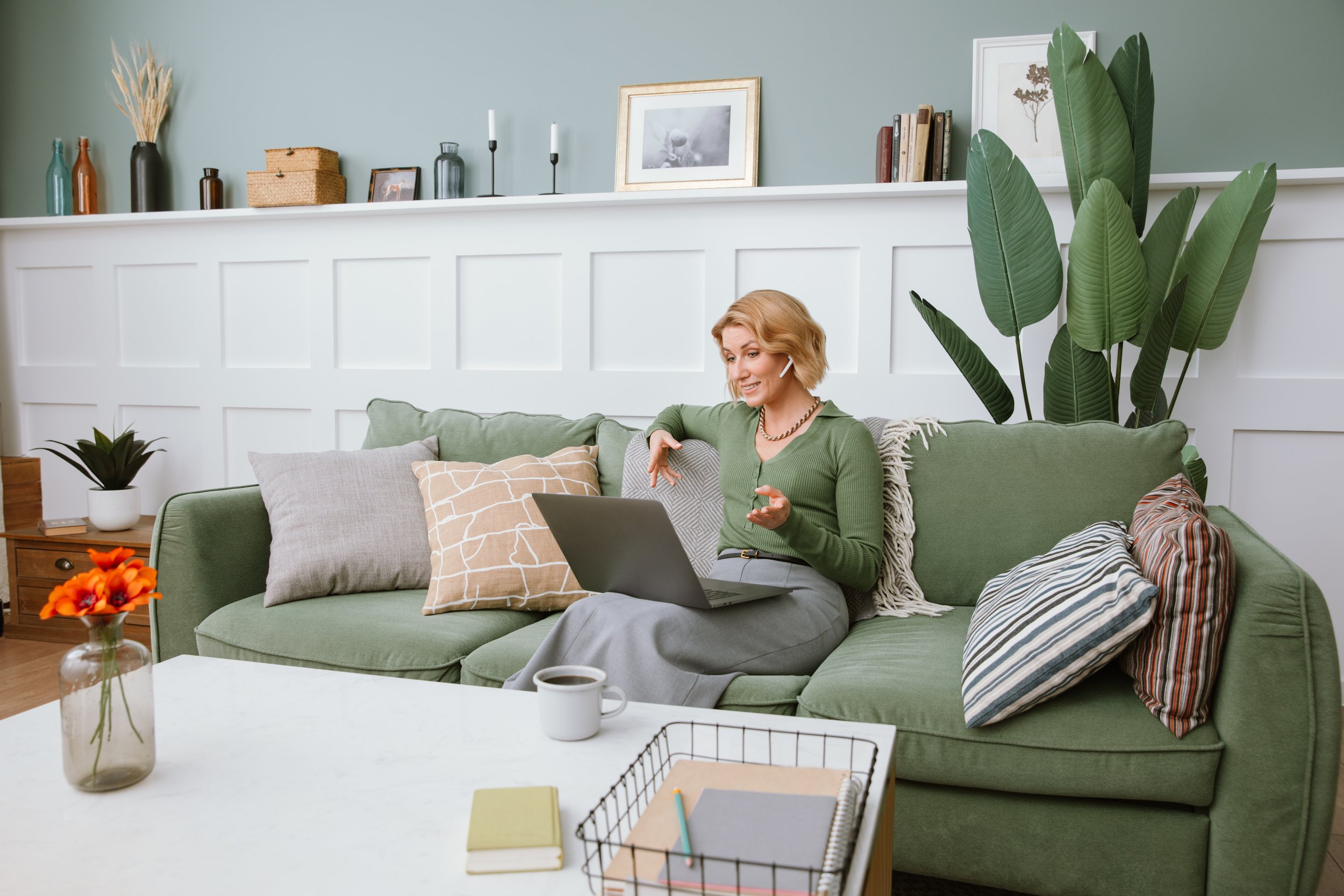 A woman sitting on a green sofa, talking on a headset, using a laptop, with cushions and a large potted plant behind her in a cozy living room.