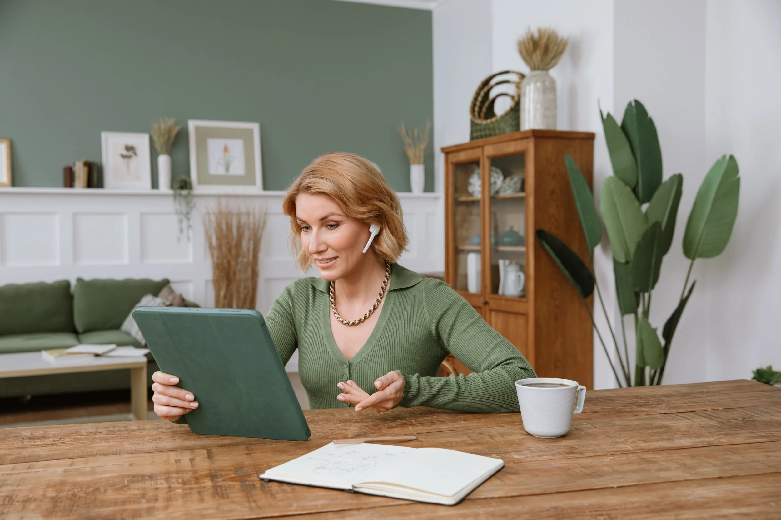 Woman wearing a green sweater and a wireless earpiece, sitting at a wooden dining table with a tablet, a notebook, a pen, and a cup of coffee or tea, in a cozy living room with green and white walls, books, plants, and decorative items.