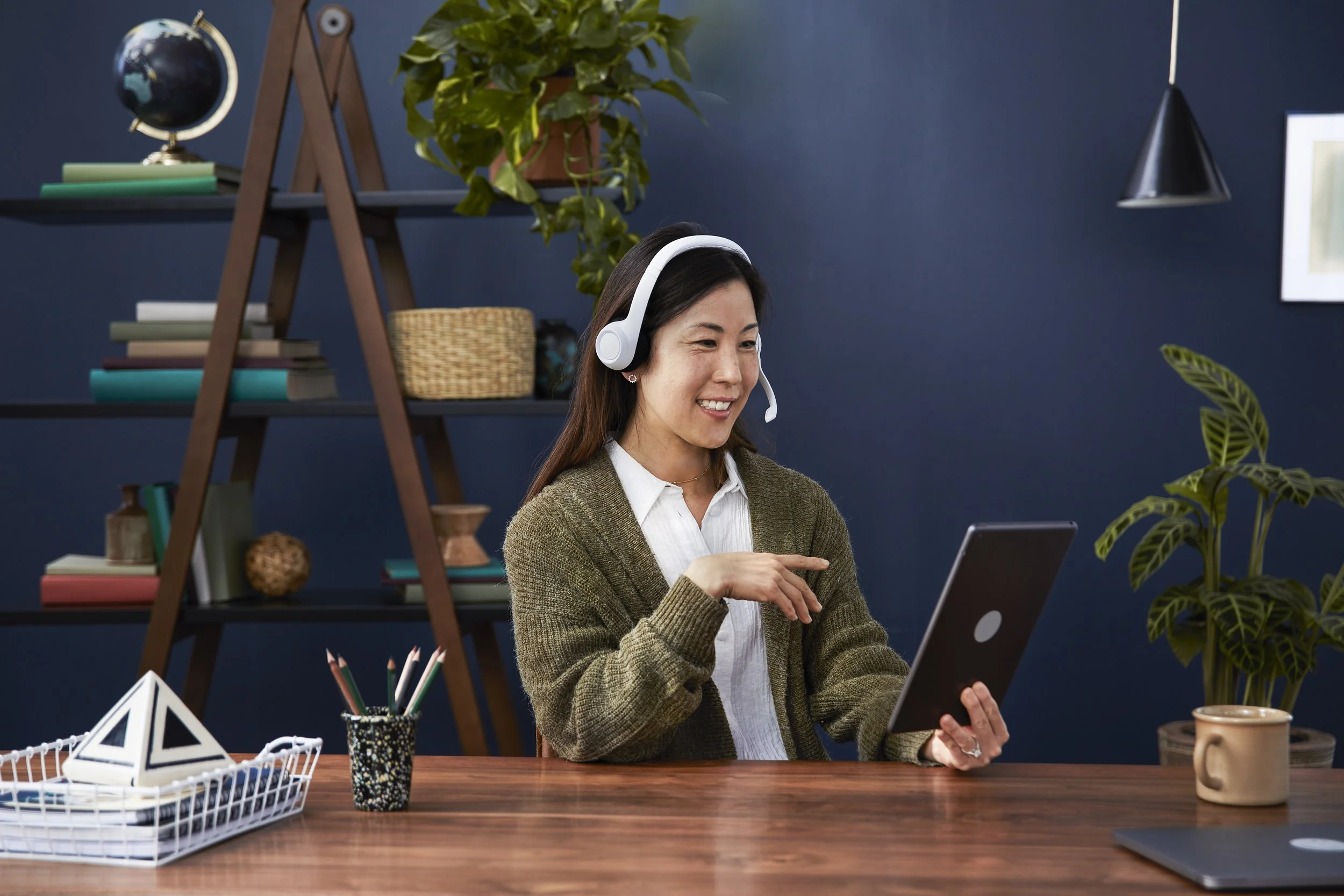 A woman with long dark hair wearing headphones, a white shirt, and a brown sweater, sitting at a wooden desk and using a tablet in a modern home office with blue walls, books, plants, and decorative items.