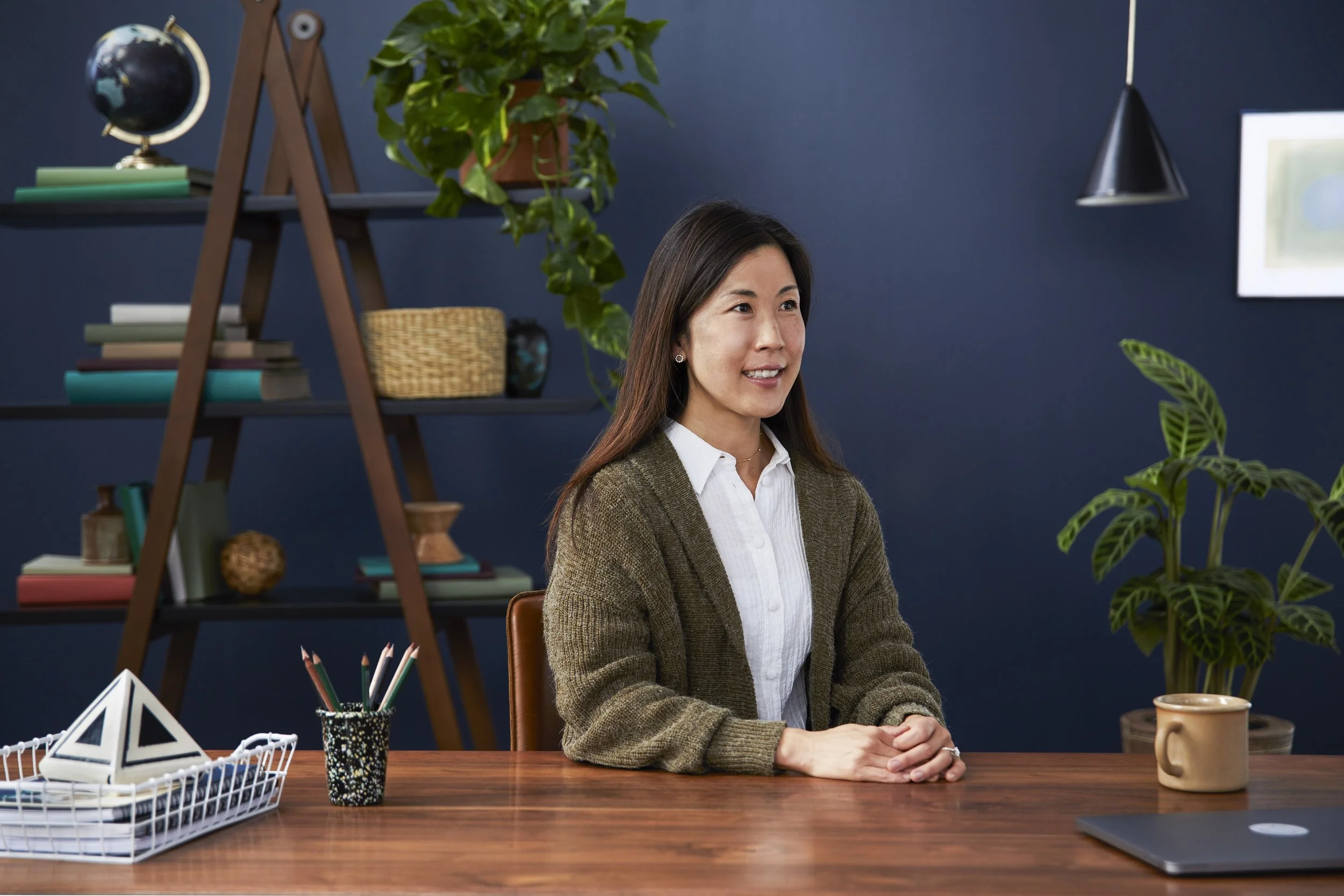 A woman sitting at a wooden desk with a beige sweater, white shirt, and dark hair, smiling, in a room with dark blue walls and shelves filled with books, plants, and decorative items.