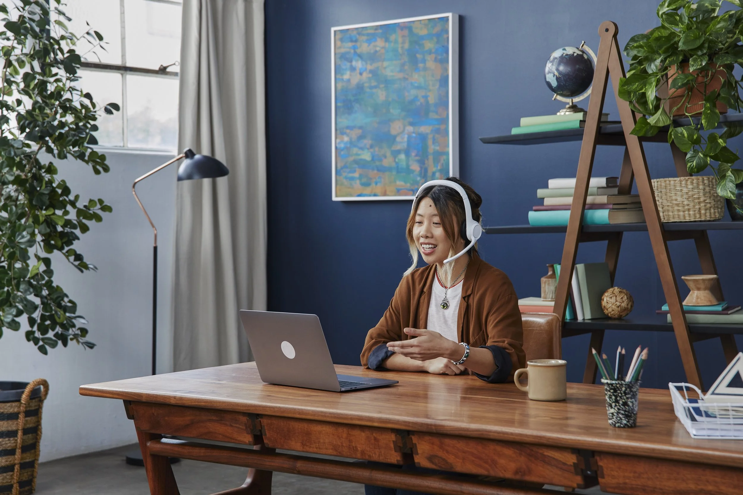A woman wearing headphones sitting at a wooden desk, smiling while looking at a laptop, in a room with blue walls, a large window, and a bookshelf with plants and decor.