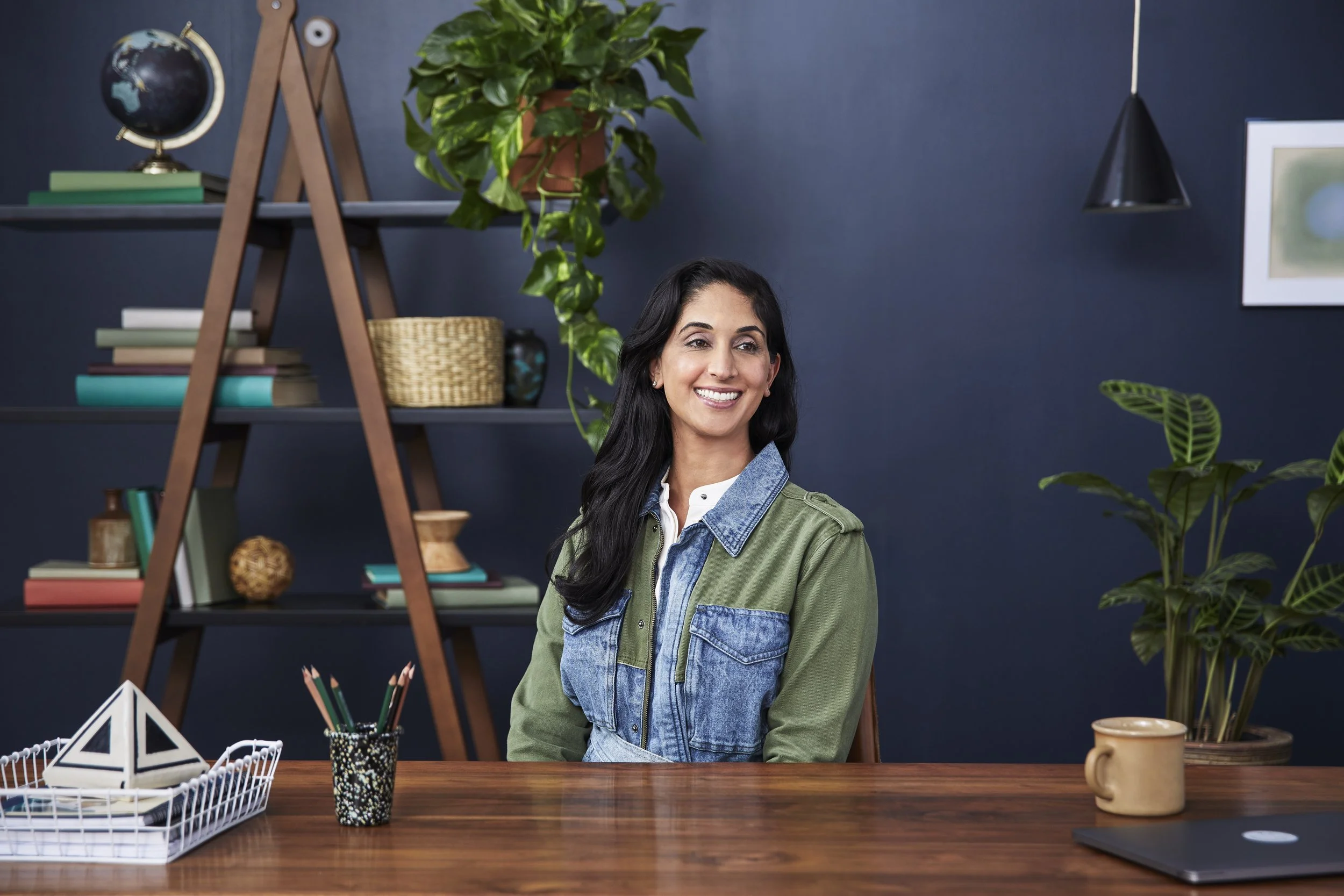 A woman with long dark hair smiling, seated at a wooden table in a home office or studio with dark blue walls, surrounded by plants, books, and decorative items.