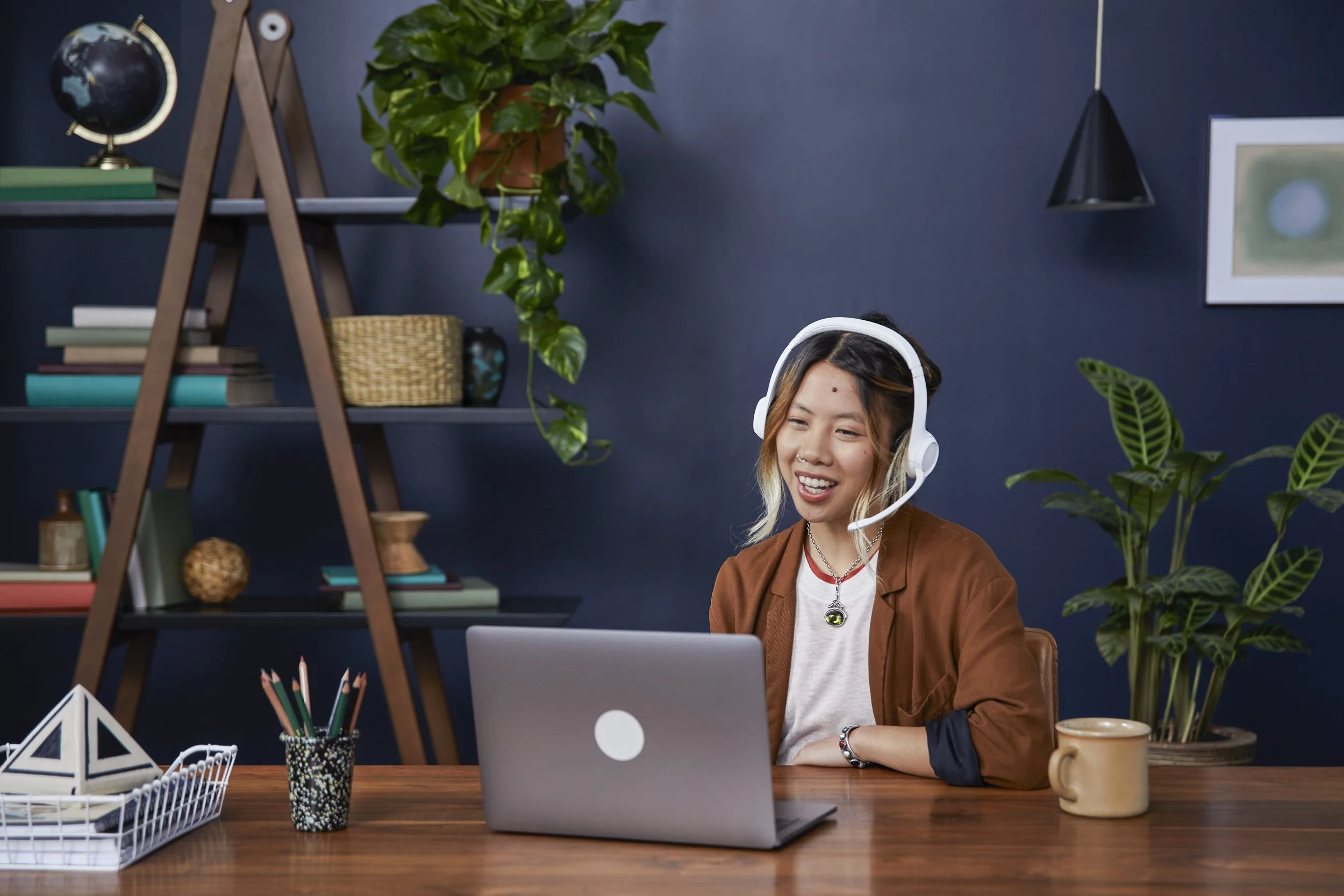 A woman with a headset sitting at a wooden desk with a laptop, plants, and stationery in a home office setting.