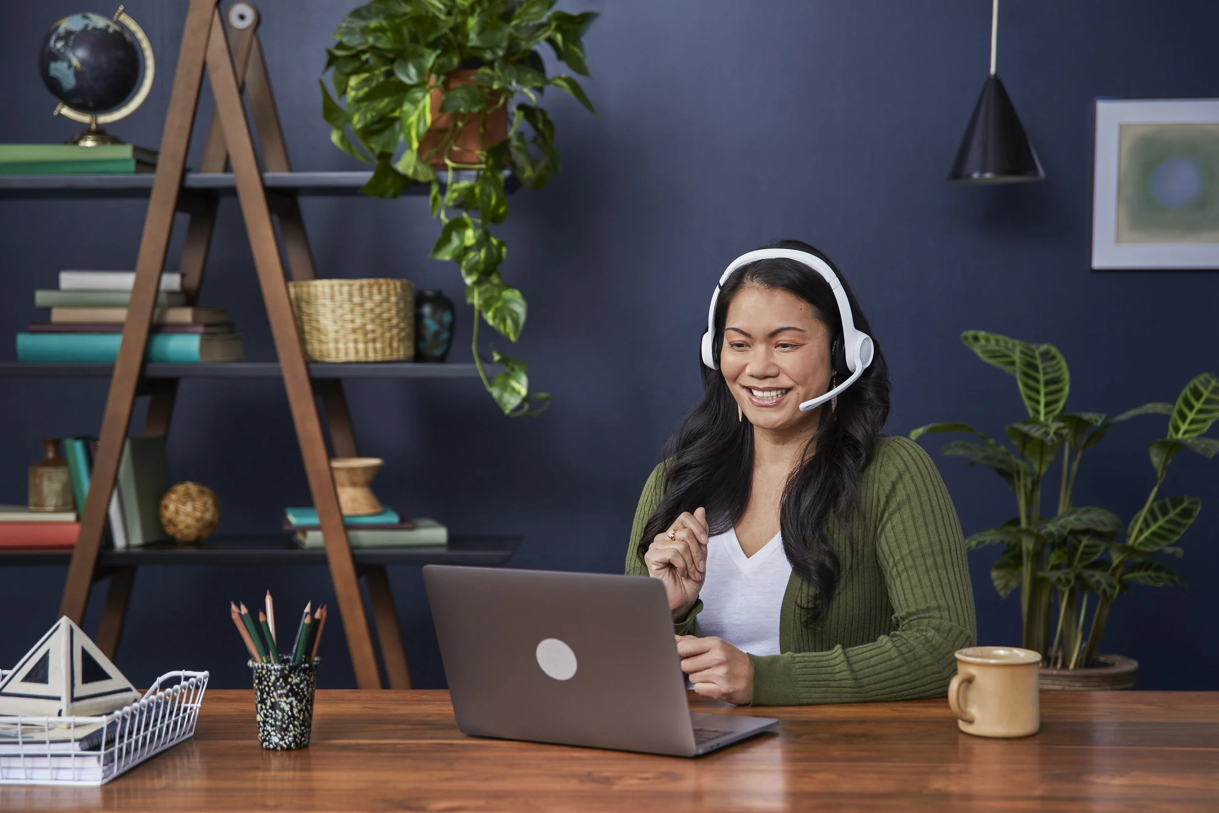 A woman with long black hair working on a laptop at a wooden desk, wearing a white shirt and green cardigan, with a headset on, in a home office decorated with plants, books, and framed artwork.