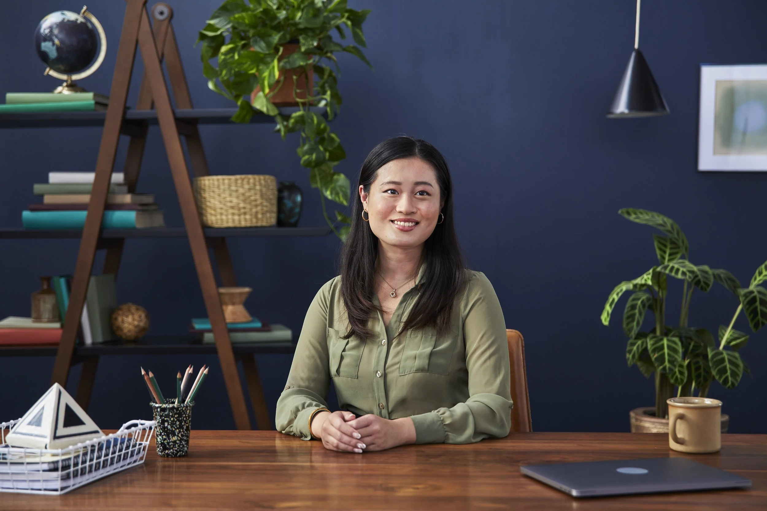 A woman with long dark hair and a green blouse sitting at a wooden desk with her hands clasped, smiling at the camera. Behind her are a dark blue wall, a shelf with books, a globe, and decorative items, along with potted plants and framed art.