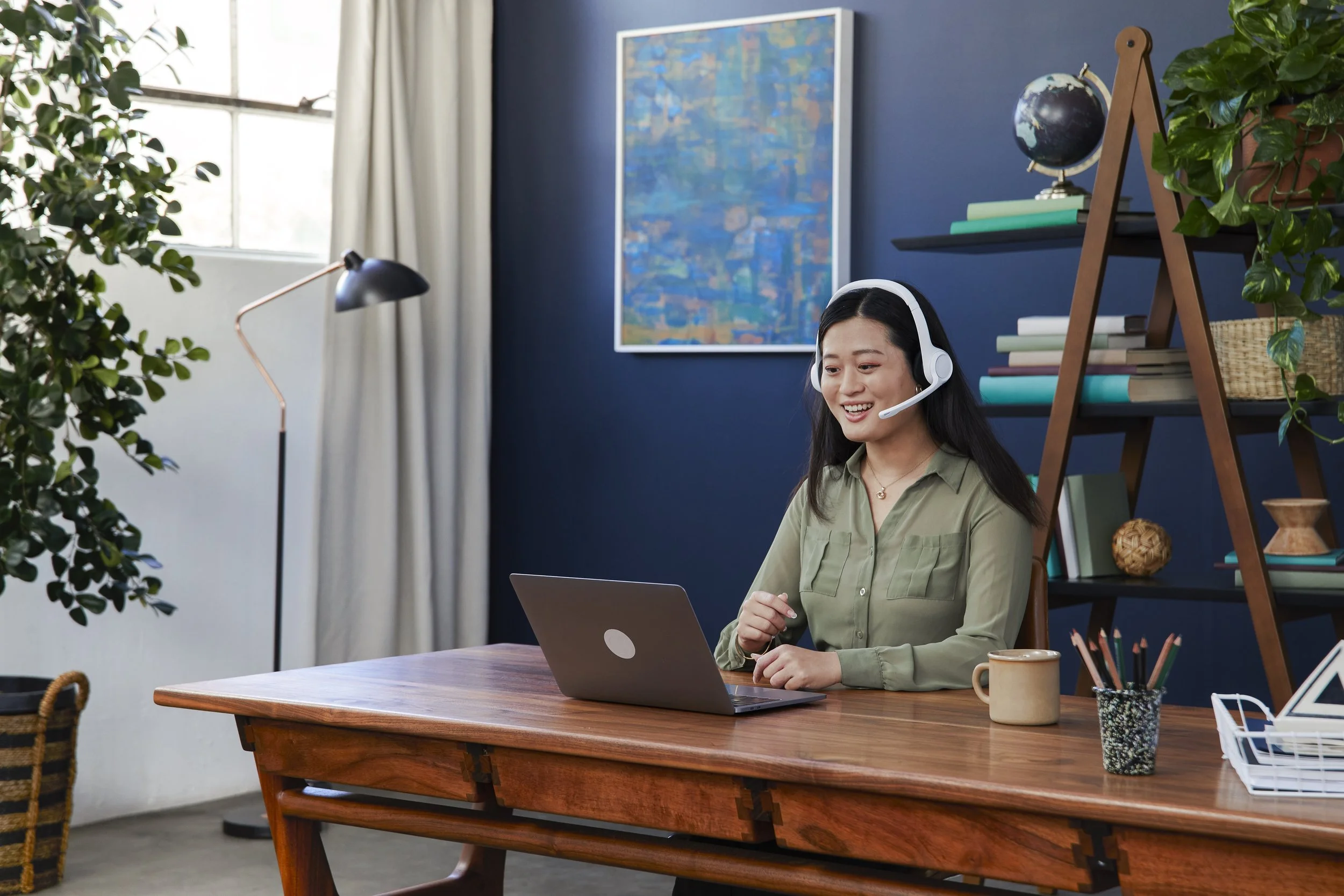 A woman wearing a headset sitting at a wooden desk with a laptop, in a well-lit room with a blue accent wall, bookshelf, plants, and artwork.