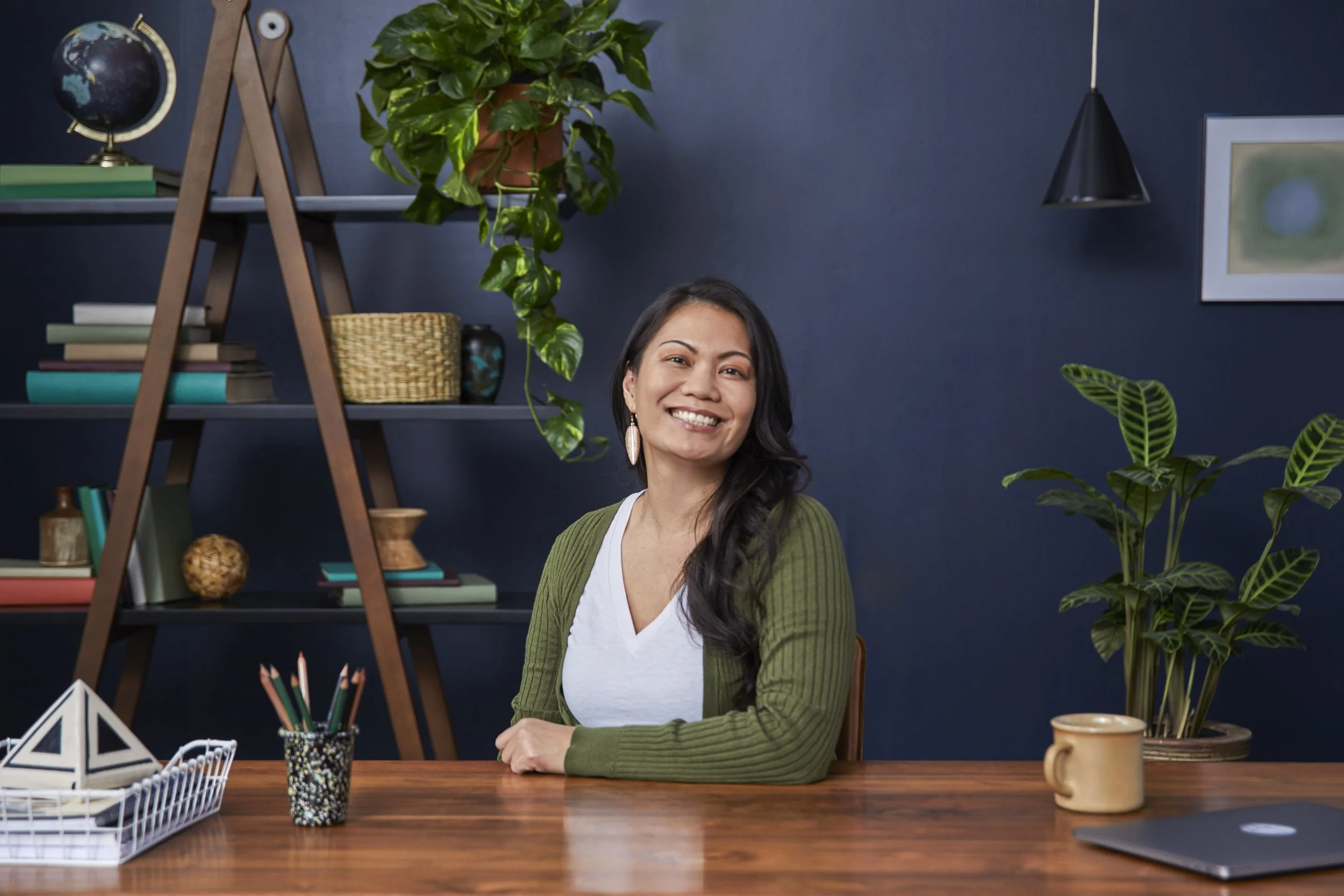 A woman with long dark hair, wearing a white shirt and green cardigan, smiling and sitting at a wooden desk with decorative items, plants, and books, against a dark blue wall with framed artwork and a hanging lamp.