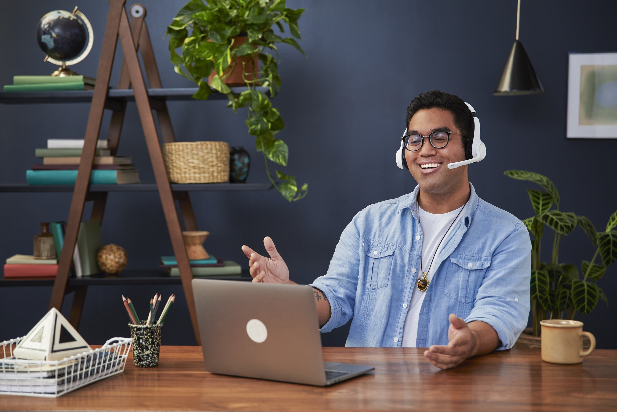 A young man with glasses and a headset sitting at a wooden desk, smiling, in a room with bookshelves, plants, and framed artwork on the walls.