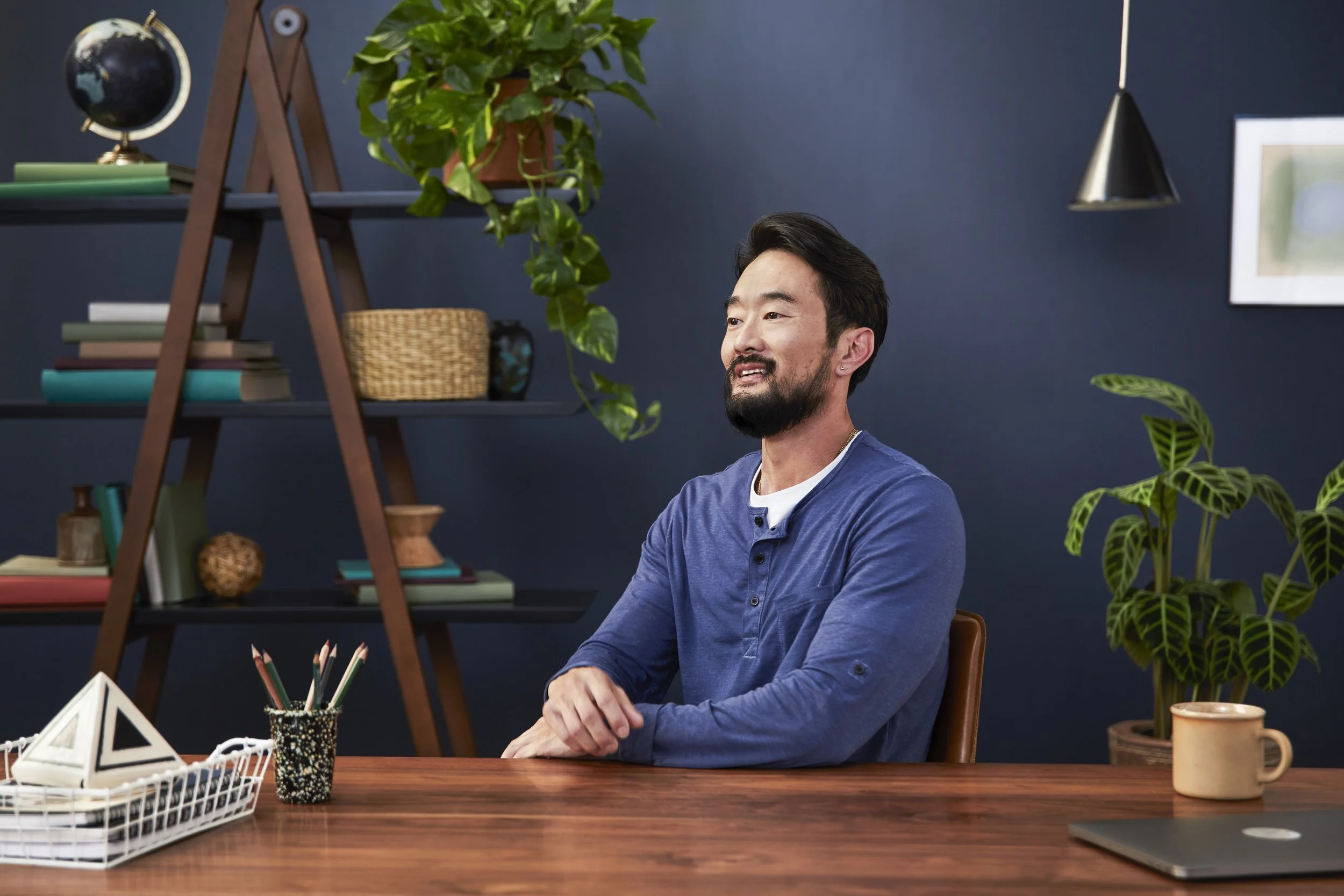A man with dark hair and a beard, wearing a blue shirt, sitting at a wooden desk in a room decorated with plants and books, with a dark blue wall and a hanging lamp in the background.