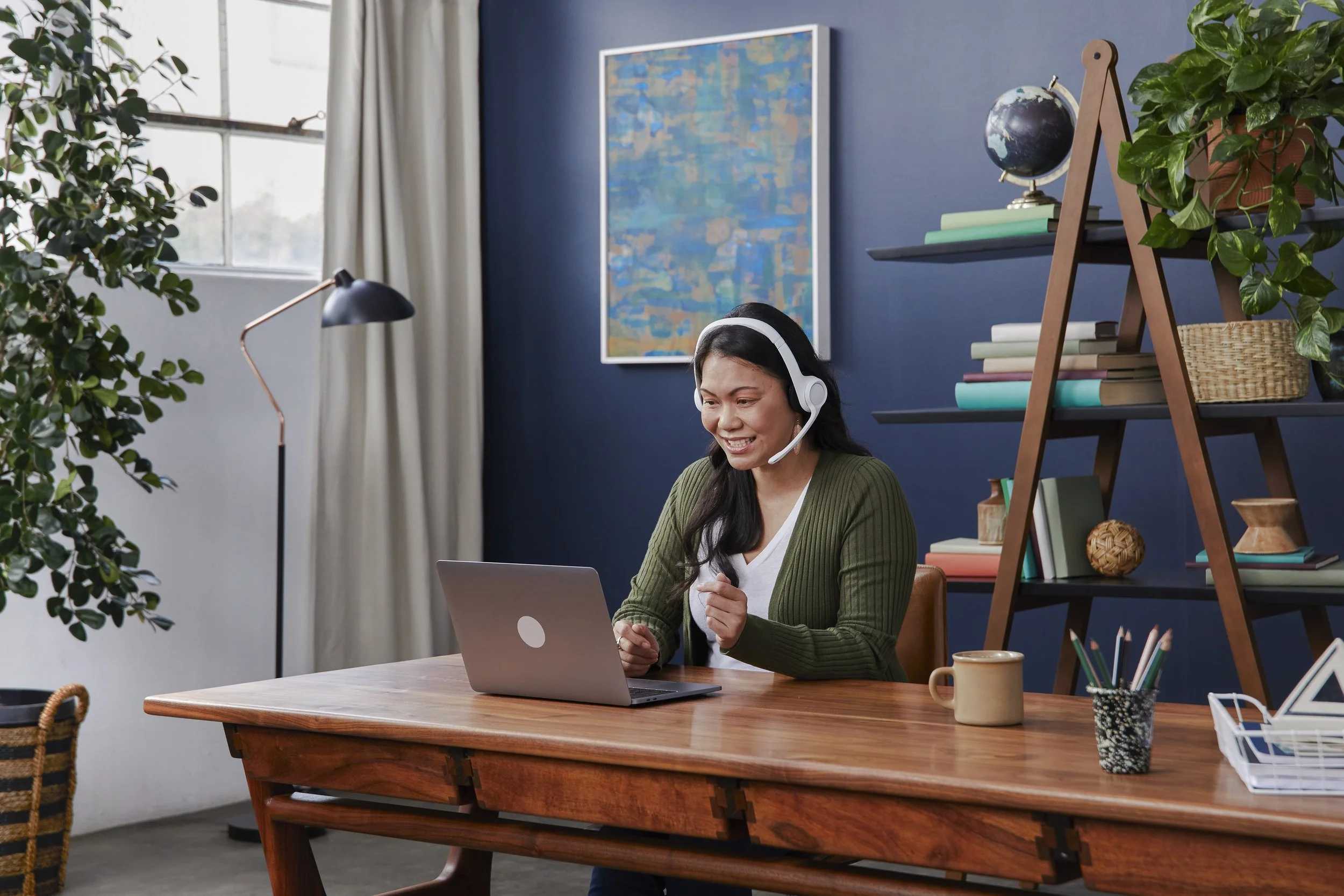 Woman with long dark hair smiling and wearing a headset, sitting at a wooden desk with a laptop computer in a well-lit, colorful office space with plants, books, and artwork on the walls.
